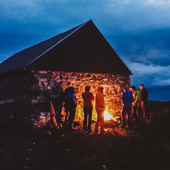 Gathering Hikers gathered around a campfire outside a remote bothy in the Scottish Highlands at dusk.
